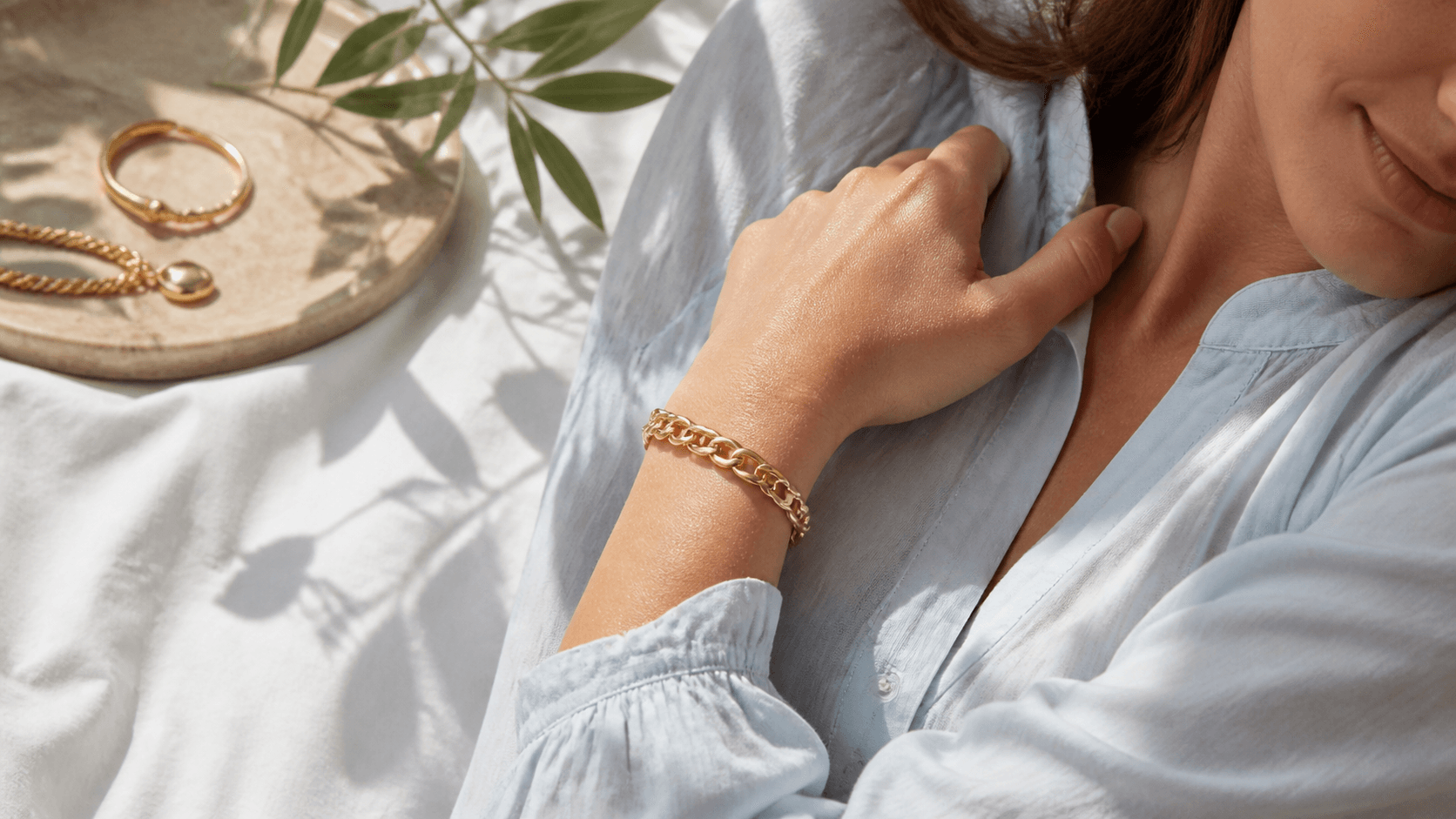 Gold heritage bracelet resting on pale linen beside sunlit white flowers and a woven straw hat.